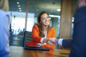 a young graduate sits across the table from her interview panel full of confidence and positivity energy . She is holding her cv and smiling at the interview panel before her and shakes hands with the new boss.She is wearing blue trousers with an orange suit jacket . in the foreground we can see the back of two of the panel , a man and a woman.