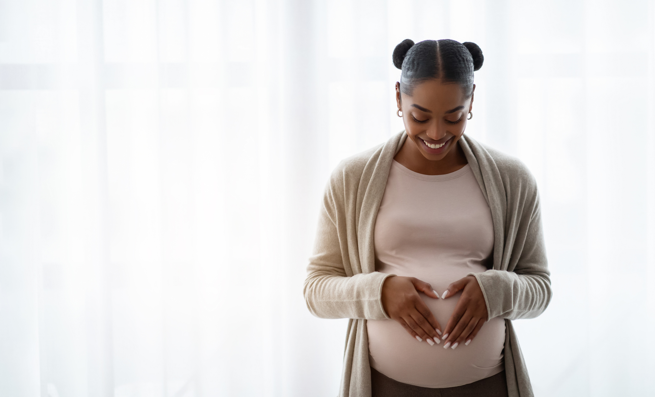 Smiling pregnant woman wearing comfy clothing holding hands on her big belly, enjoying her pregnancy, expecting baby, standing by window at home, panorama with copy space