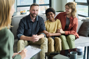 family sitting on couch during counseling session, smiling and holding hands. Professional counselor in foreground with clipboard in hand and tablet on table