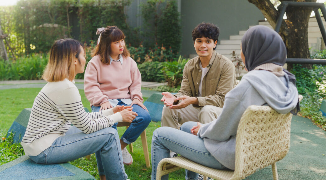 Small group of people sit in a close circle and talk to a therapist in park. Smile people sharing story happy speak diverse people sitting in circle at group therapy session with psychologist.